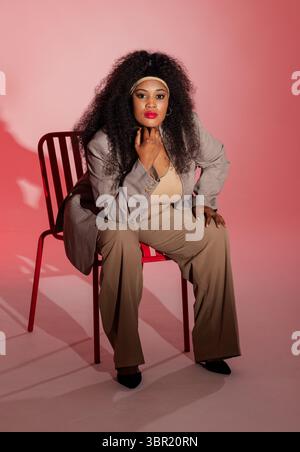 Donna afroamericana seduta in studio su una sedia di metallo rossa che indossa un blazer appoggiato toccando il mento. Professionale, sicuro, elegante, elegante, pronto, c Foto Stock