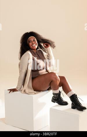 Donna afroamericana seduta su cubetti in studio, che solleva capelli ricci in cardigan e stivali da combattimento. Ritratto, diversità, moda, minimalista, casual, Foto Stock