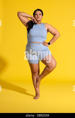 Donna asiatica in equilibrio sulla gamba destra con sfondo giallo in studio che indossa smartwatch e orecchini. Atletica, fitness, minimalista, vitalità, benessere, att Foto Stock