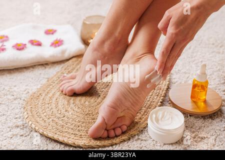 Mani femminili che applicano crema idratante ai tacchi con olio e asciugamano, cure termali a casa per piedi sani e morbidi, bellezza cura della pelle routine Foto Stock