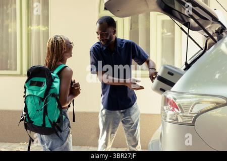 Uomo e donna di colore che caricano i bagagli nell'auto e si preparano per il viaggio in auto, circondati da valigie e sole estivo nel vialetto. Avventura gioiosa con divertimento e viaggi di piacere insieme. Foto Stock