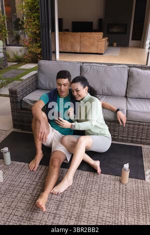 Diverse coppie sedute sul tappetino da palestra del patio accanto al divano in rattan, controllando smartphone e bottiglie d'acqua. Esterno, relax, moderno, tempo libero, stile di vita, c Foto Stock