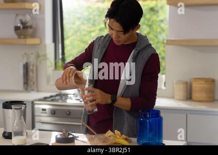 Uomo asiatico che versa frullato di cioccolato dallo shaker al bicchiere in cucina con contenitore supplementare Foto Stock