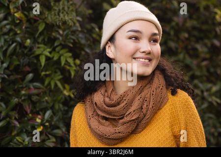 Sorridendo e guardando bene, donna asiatica in piedi accanto alla siepe nel parco con maglione, sciarpa, berretto Foto Stock