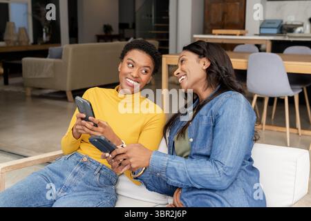Diverse amiche che ridono mentre condividono gli schermi dello smartphone sul divano nel soggiorno open space Foto Stock
