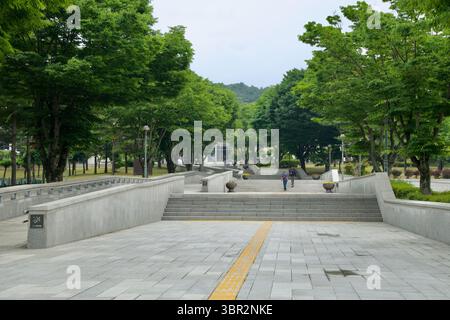 Gongju, Corea del Sud - 13 giugno 2025: Una passerella in pietra con pavimentazione tattile e gradini conduce attraverso alberi frondosi verso l'ingresso in vetro di Gongju nati Foto Stock