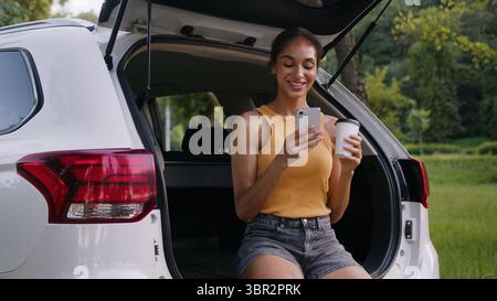Donna afroamericana che beve caffè sorridente rilassante ragazza felice che tiene cellulare smartphone social media gadget auto auto auto Foto Stock