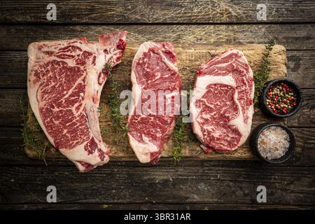 Varietà di bistecche fresche di carne cruda Black Angus prime T-Bone, New York, Ribeye e stagionatura su sfondo di legno, vista dall'alto Foto Stock