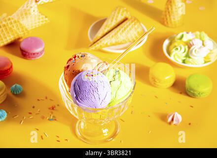 Vari cucchiai di gelato colorati o piatti di palle con coni per waffle, macaron su sfondo giallo Foto Stock