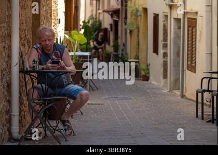 Chania, Creta, Grecia - 22 giugno 2025: Uomo seduto ad un tavolo in un vicolo stretto di Chania mentre usa il suo telefono. Foto Stock
