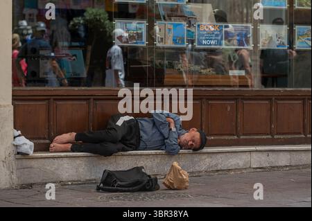 Chania, Creta, Grecia - 22 giugno 2025: Un uomo giace addormentato su una panchina di pietra fuori da un'agenzia di viaggi a Chania, Creta, in mezzo ai turisti. Foto Stock
