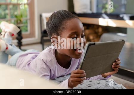 Bambina afroamericana sdraiata sul cuscino in soggiorno tenendo un tablet vicino alla luce della finestra Foto Stock