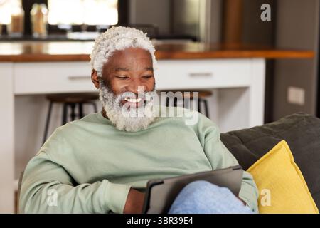 Uomo afroamericano anziano seduto su un divano grigio con cuscino che tiene il tablet vicino al bancone della cucina Foto Stock