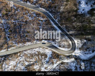 Veduta aerea di una tortuosa strada asfaltata si snoda attraverso un paesaggio dove macchie di neve che si scioglie contrastano con il marrone e il verde degli alberi, con un'auto solitaria che naviga lungo la curva, Zagabria, Croazia Foto Stock