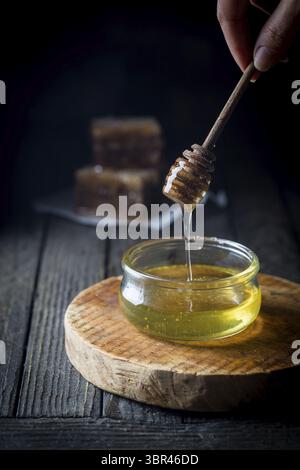 Miele che gocciola dal cucchiaio di miele in un vaso di vetro con favo a nido d'ape su sfondo scuro Foto Stock