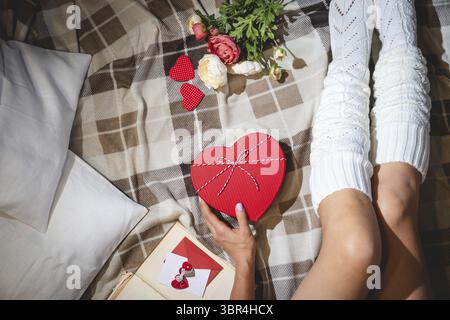San Valentino festa piatto posare con regalo in scatola a forma di cuore e le gambe in calze bianche su fondo letto o pavimento, vista dall'alto Foto Stock