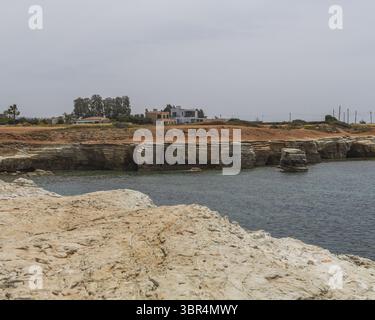 View of the rugged coastline where the tan earth meets the tranquil turquoise sea under a muted sky, a home nestled amidst the landscape, Paphos, Paph Foto Stock