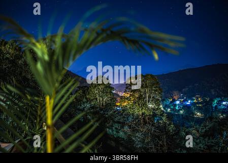 Luci del villaggio e montagne incorniciate da Palm Leaf sotto Starry Sky a Itaipava, Petrópolis, Brasile Foto Stock