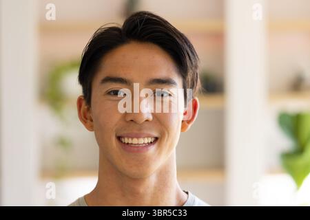 Uomo asiatico sorridente calorosamente in casa ufficio con scaffali in legno sfocati e piante verdi Foto Stock