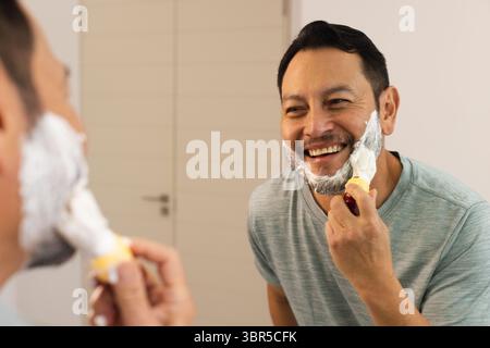 Uomo asiatico che indossa una t-shirt verde chiaro radersi al lavandino del bagno usando rasoio e schiuma nello specchio Foto Stock