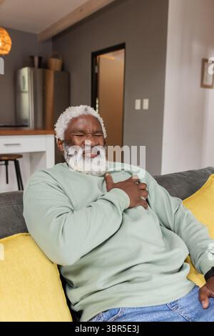 Senior afroamericano che stringe il petto seduto su un divano grigio nell'area soggiorno con cucina aperta Foto Stock