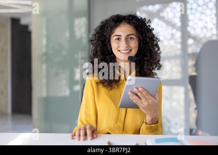 Una donna sorridente con i capelli ricci che indossa una cuffia e tiene in mano un tablet, guarda direttamente la fotocamera in ufficio. Foto Stock