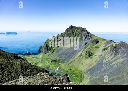 Vista delle aspre scogliere verdi che si tuffano nel mare blu, con una piccola pista di atterraggio annidata nella valle sottostante, Vestmannaeyjabær, Vestmannaeyjabær, Islanda. Foto Stock