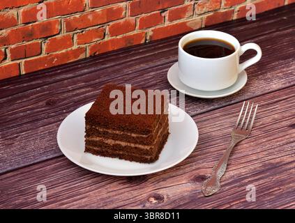 Un pezzo di torta a strati di cioccolato su un piatto, una forchetta e una tazza di caffè nero caldo su un tavolo di legno. Primo piano. Foto Stock