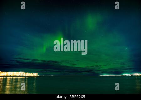 Vista delle vivaci danze dell'aurora boreale attraverso il cielo notturno, che proiettano un bagliore etereo sulle acque tranquille e sulle luci lontane della città, Reykjavík, Reykjavíkurborg, Islanda. Foto Stock
