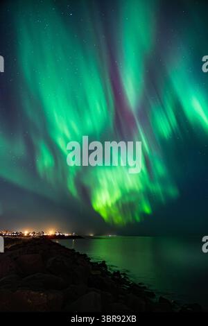 Veduta aerea di una radiosa aurora boreale danzante nel cielo notturno, che getta eteree sfumature di verde e viola sulle tranquille acque della costa, l'Islanda. Foto Stock