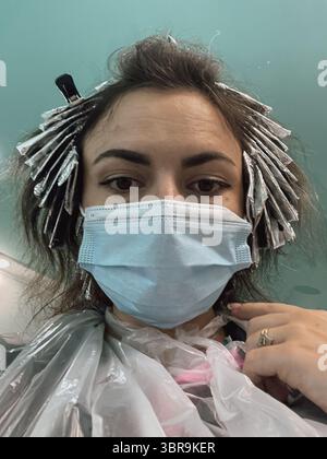 Ragazza che prende un selfie mentre tinge i capelli con una maschera medica su Foto Stock