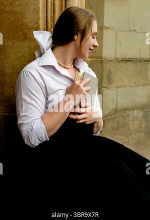 Una giovane donna con una camicia bianca e una collana di perle tiene in mano un libro, appoggiata contro un muro di pietra. Foto Stock