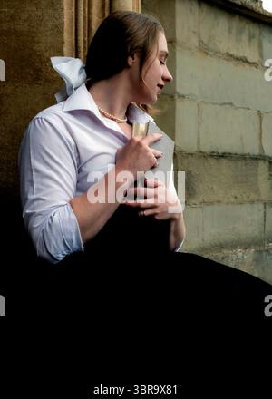 Una donna con una camicia bianca e una gonna nera tiene un libro vicino al suo petto, appoggiato contro un muro di pietra. Foto Stock