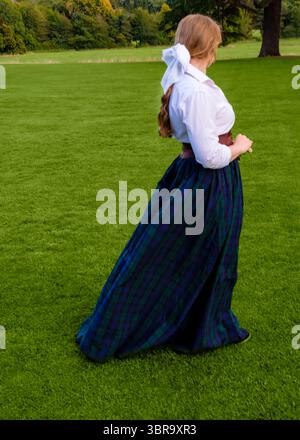 Una donna con una camicia bianca, una cintura marrone e una lunga gonna a quadri blu e verde si staglia su un campo erboso. Foto Stock