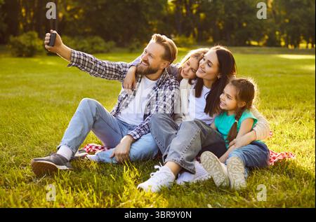 Famiglia felice seduta insieme sull'erba verde estiva a scattare selfie utilizzando il cellulare abbracciando la condivisione Foto Stock