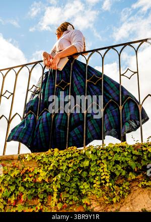 Una donna con una camicia bianca e una gonna a quadri si appoggia a una ringhiera in ferro battuto, tenendo in mano un libro. Il cielo è blu con alcune nuvole. Foto Stock