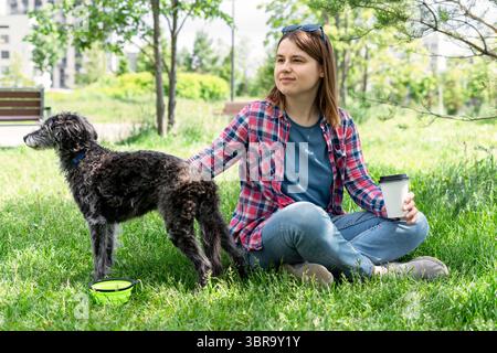 Giovane donna seduta sull'erba con un soffice cane anziano grigio di razza mista, bedlington terrier Whiippet, gustando un caffè in un parco soleggiato, circondato da greene Foto Stock