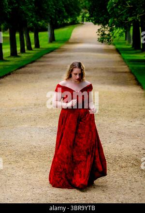 Una donna con un vestito rosso cammina lungo un sentiero fiancheggiato da alberi. Foto Stock