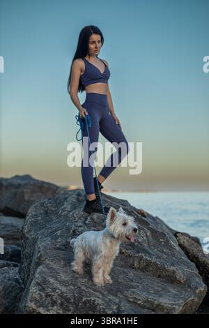 Una bella donna bruna che indossa un abbigliamento da fitness si erge su una roccia vicino alla spiaggia e si gode una tranquilla passeggiata al tramonto con il suo cane bianco accanto a lei, mostrando la sua pelle liscia dopo l'epilazione Foto Stock