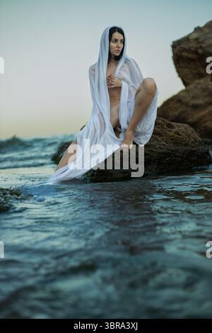 La bella figura bruna, drappeggiata in tessuto puro, si trova graziosamente su una roccia vicino al mare che il sole tramonta dietro di lei, gettando un sereno bagliore sulla superficie dell'acqua Foto Stock