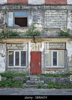 Edificio abbandonato in rovina con vernice sbucciata, intonaco sbriciolato e crepe. Arbusti e erbe ricoperte da muri trascurati e travi intemperie. Foto Stock