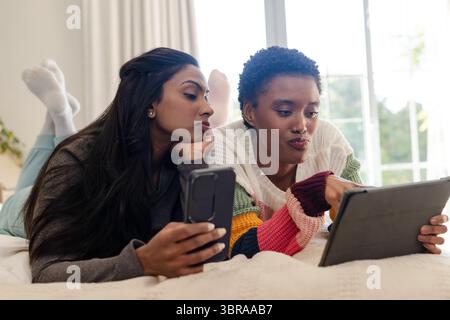Diverse amiche distese a letto in camera da letto utilizzando smartphone e tablet su biancheria da letto bianca Foto Stock