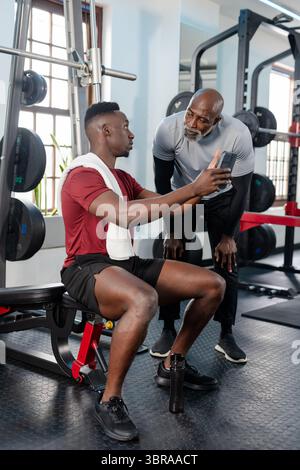 Allenatore e apprendista afroamericano che si appoggia sullo smartphone sulla panca per pesi con asciugamano in palestra Foto Stock