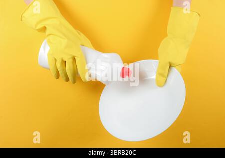Le mani femminili con guanti in gomma gialla trattengono il flacone di detergente e la piastra bianca su sfondo giallo studio. Concetto di lavastoviglie Foto Stock