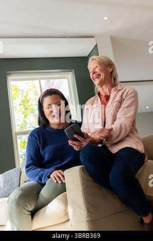 Diverse amiche che ridono e condividono lo smartphone mentre si siedono sul divano beige nel soggiorno Foto Stock