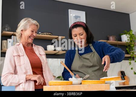 Diverse amiche si mescolano e ridono sulla pastella per torte con la spatola sul ripiano in cucina Foto Stock
