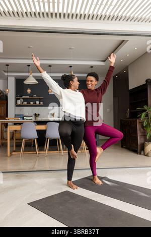 Diverse amiche che praticano posa sugli alberi su tappetini neri da yoga nel soggiorno con luci a sospensione Foto Stock
