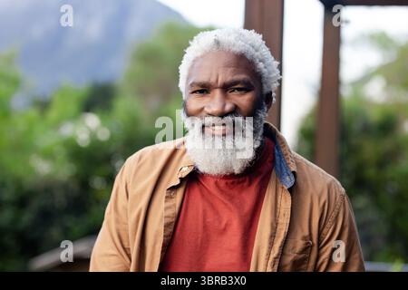 Uomo afroamericano senior che indossa una giacca marrone camicia rossa in piedi nel cortile con pergola e montagna Foto Stock