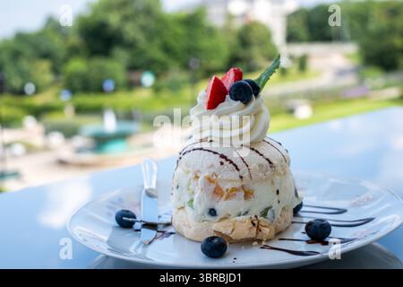Torta gelato a strati adornata con panna montata, fragole e mirtilli, presentata su un piatto bianco, con una splendida vista all'aperto nel bac Foto Stock