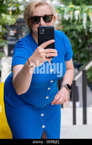 Una donna in abito blu cattura selfie all'aperto, circondata da una vegetazione lussureggiante, che irradia fiducia e abbraccia il benessere in un ambiente sereno e vivace Foto Stock